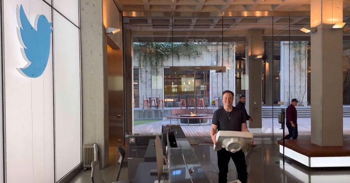 Elon Musk, wearing a black shirt and black pants, is carrying a large sink and smiling as he enters Twitter's California headquarters. He is passing by a desk in front of a wall bearing Twitter's blue bird icon.