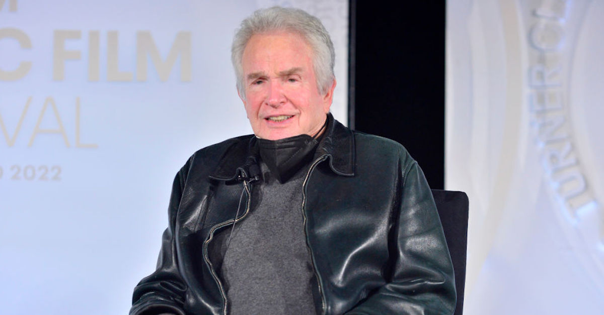 Warren Beatty is wearing a black leather jacket, a gray shirt, and a black mask around his neck. He is sitting for an interview and appears to be looking at an audience. He is white withe gray hair.