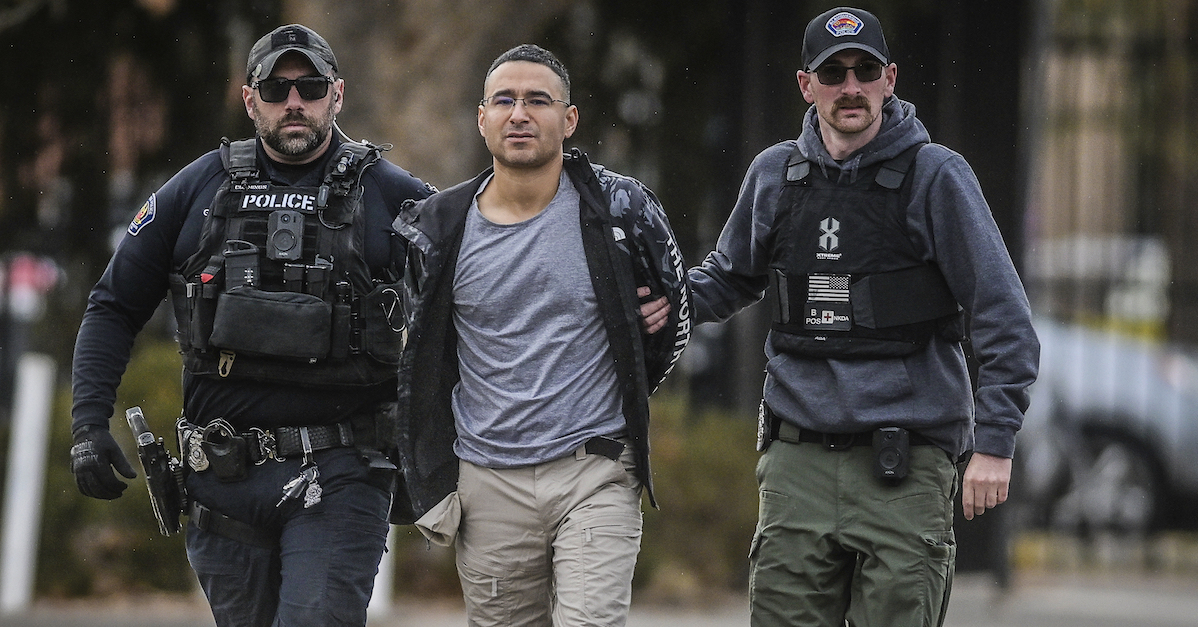 Solomon Pena, wearing a blue T-shirt, khaki cargo pants, and an unzipped jacket, is flanked by two police officers wearing tactical vests over their clothes.